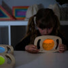 Una pequeña niña sentada en una mesa con un juguete de madera, un holograma de Paul Feeley, presentado en dribble, arte interactivo, bioluminiscencia, luz parpadeante, iluminación volumétrica.