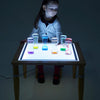Laden Sie das Bild in den Galerie-Viewer, Una niña sentada en una mesa con una superficie iluminada, un holograma de Cerith Wyn Evans, presentado en la Sociedad de CG, arte interactivo, bioluminiscencia, luminiscencia, arte académico.